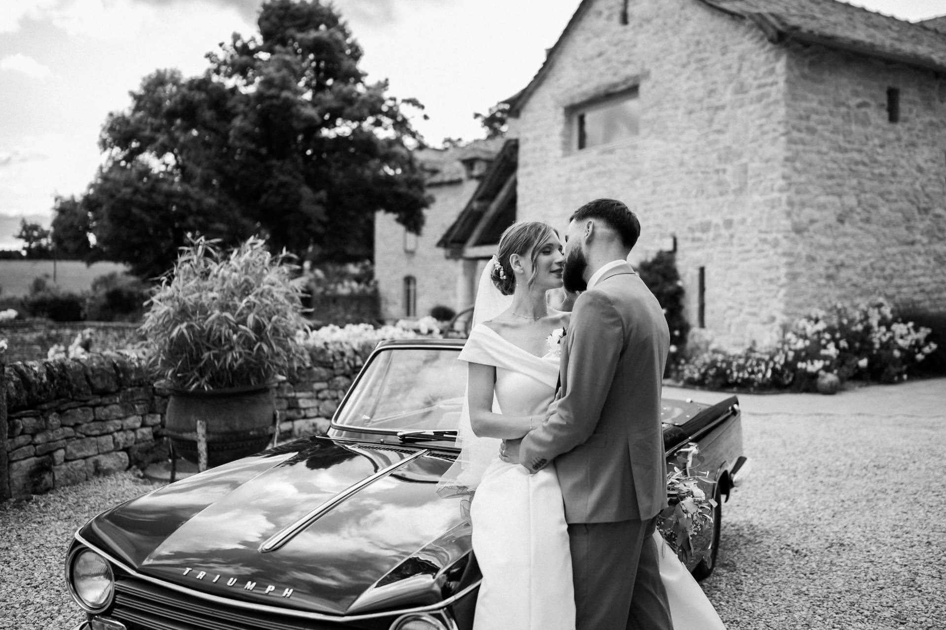 Couple de mariés enlacé devant une voiture ancienne, en noir et blanc
